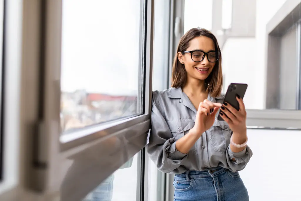 Smiling woman in glasses checking her phone by a window