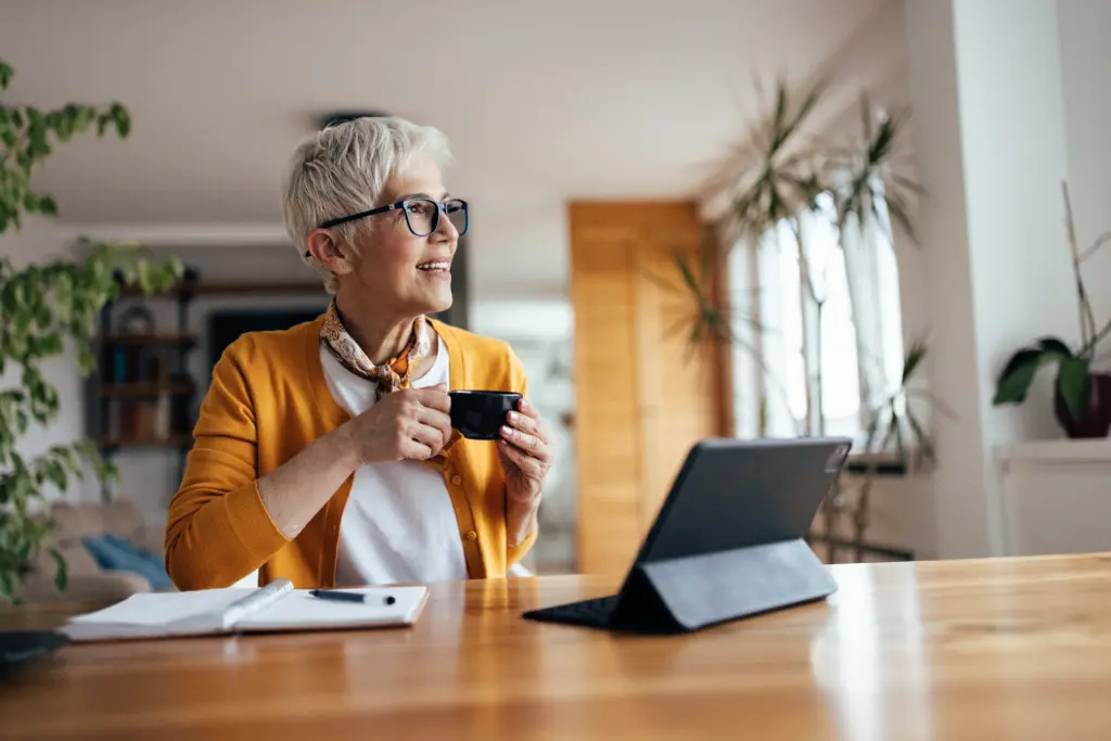 Smiling senior woman with a coffee mug and a tablet at her kitchen table